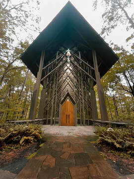 A photo of Anthony Chapel, a modern glass and wood chapel situated in the Garvan Woodland Gardens in Hot Springs, Arkansas. The chapel has a soaring vaulted glass ceiling and floor-to-ceiling windows, which provide stunning views of the surrounding forest. The chapel is surrounded by trees and is located on a hillside overlooking Lake Hamilton.  T